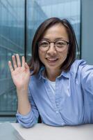 An Asian businesswoman wearing glasses smiles and waves during a call. The setting is a modern office, conveying a sense of communication and friendly interaction. photo