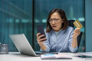 Asian businesswoman in office experiencing confusion during online transaction. Holding smartphone and credit card with concerned expression, signifying possible payment problem. photo
