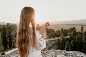 Happy woman in white boho dress on sunset in mountains. Romantic woman with long hair standing with her back on the sunset in nature in summer with open hands. Silhouette. Nature. Sunset. photo