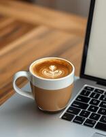 Coffee cup with latte art placed beside a laptop on a wooden table during the day photo