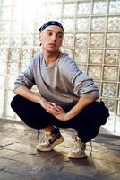 A man in a gray sweatshirt and black pants sits in a squat with his hands clasped in his lap, looking to the side, in front of a glass block wall photo