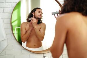 A man with curly hair grooming himself in the bathroom while looking into a round mirror photo