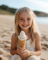 A joyful child holds a cone of ice cream while seated on the sandy beach during a warm afternoon photo