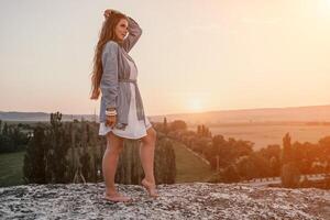 Happy woman standing with her back on the sunset in nature in summer with open hands. Romantic beautiful bride in white boho dress posing with mountains on sunset photo