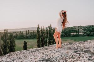 Happy woman in white boho dress on sunset in mountains. Romantic woman with long hair standing with her back on the sunset in nature in summer with open hands. Silhouette. Nature. Sunset. photo