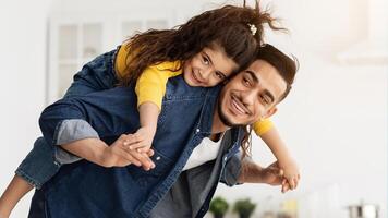 Cheerful arab dad carrying his cute little daughter on back and laughing, happy middle eastern family playing and having fun together at home, joyful female child enjoying spending time with daddy photo