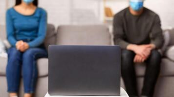 Watching News. Selective focus on personal computer, spouses in surgical masks sitting on the couch in blurred background photo