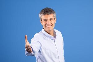 A middle aged man in his 50s stands against a solid blue background, smiling and extending his right hand in a welcoming gesture. His attire is casual, reflecting a friendly lifestyle. photo