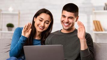 Happy lovely couple making call on laptop in living room, waving at screen, chatting with friends and family photo