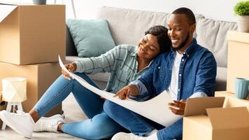 Black Spouses Planning Home Renovation, Checking Blueprints And Color Palette While Sitting Among Cardboard Boxes In Their New Apartment, African American Couple Decorating House After Moving photo
