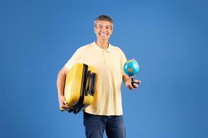 A middle aged European man stands confidently against a blue backdrop, holding a yellow suitcase in one hand and a globe in the other, embodying a spirit of adventure and exploration. photo