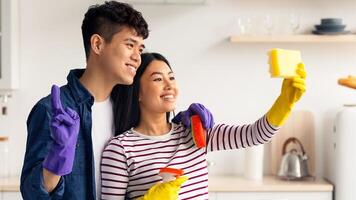 Loving asian couple taking selfie while cleaning kitchen together, using sponge as smartphone, hugging and smiling, young man and woman having fun while house-keeping at home, side view photo