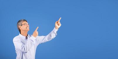 A mature man in his 50s gestures with both hands, pointing upward at copy space with a big smile. The clear blue background enhances his lively expression, suggesting excitement or celebration. photo