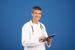 A mature Caucasian doctor in his 50s, wearing a white coat and stethoscope, smiles while using a tablet against a solid blue background. He appears engaged and ready for medical consultations. photo