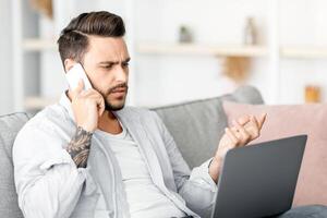 Discontented millennial man talking on phone and looking at laptop, having problem with internet connection, sitting at home, copy space. Displeased customer calling to hotline service photo