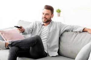 Leisure and entertainment. Happy guy watching TV switching television channels with remote control, sitting on sofa at home, copy space. TV programming concept photo