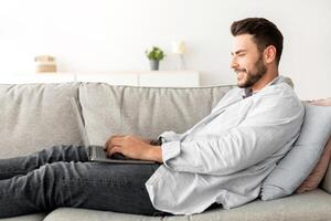 Happy millennial guy using laptop computer while resting at home on sofa, side view, free space. Excited man chatting with friends online or male freelancer working from home photo