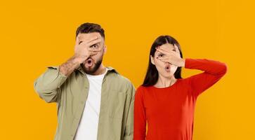 A young Caucasian couple stands together against a bright yellow background. They engage in a playful gesture, covering their faces with their hands while expressing joy. photo
