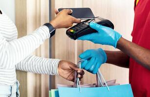 Unrecognizable Black Female Making Mobile Payment For Delivery Putting Smartphone To Terminal Receiving Shopping Bags From Deliveryman Indoors. Cropped Shot. Courier Service Concept photo