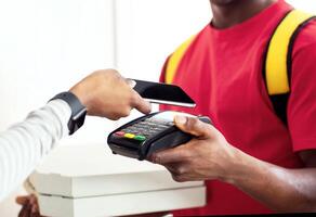 Black Courier Delivering Pizza And Receiving Payment, Customer Lady Putting Phone To Terminal Standing At Home. Credit Card In Your Smartphone, Bank Service. Cropped, Selective Focus On Cellphone photo