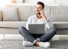 Distant work and mobile communication. Happy freelancer man talking on cellphone and using laptop while working remotely from home, sitting on floor near sofa in living room, free space photo
