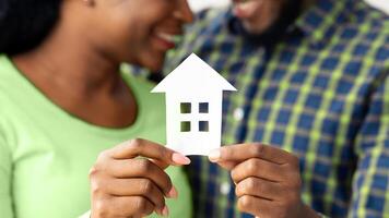 African American Spouses Holding Cutout Paper House Figure In Hands, Celebrating Buying New Property. Conceptual Photo For Family Housing, Real Estate Or Relocation, Closeup With Selective Focus