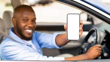 Closeup of young smiling African American man showing mobile phone with white empty screen for mock up sitting on driver seat in new luxury car, holding gadget in hand, selective focus on device photo