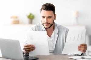 Millennial male self-entrepreneur working with papers at desk at home office, checking annual financial reports, free space. Young man reading documents, sitting at workplace with computer photo