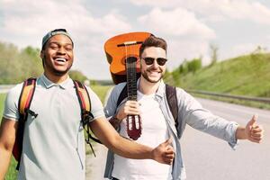 Two guys with guitar hitchhiking on road, stopping passing cars with thumb up gesture, having fun summer adventure, copy space. Multiracial friends traveling by autostop, enjoying their journey photo