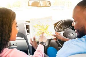 Road Trip Concept. Rear back view from passenger's seat of smiling black man and lady holding and looking at paper map sitting inside car, choosing location, woman pointing at destination point photo