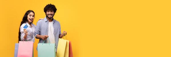 Consumerism concept. Excited indian couple with credit card and shopping bags, buying clothes remotely on yellow background. Positive guy and his girlfriend encouraging contactless payment, copy space photo