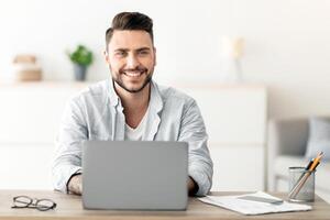 Successful freelancer. Happy man sitting at workplace with laptop computer, smiling and looking at camera, working remotely on computer at home office, copy space. Guy enjoying online distant job photo