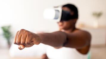 Virtual training concept. Overweight black lady in VR glasses using fighting simulator, making punch at home, focus on clenched fist. Plus size Afro woman boxing with augmented reality app photo