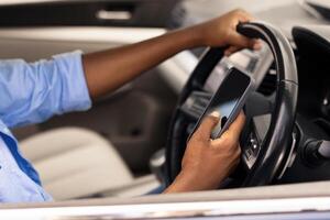 Positive black male driver holding mobile phone while driving nice auto, using new mobile application for navigation going on vacation or making summer trip, stuck in traffic jam, profile side view photo