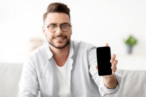 I recommend this website. Young man showing smartphone with blank screen to camera, mockup for your ad or design. Guy sitting on sofa at home, holding cellphone in hand, selective focus photo