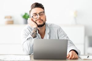 Overworked young freelancer man sitting at workplace and sleeping, napping while working on laptop computer, placed head on hand, feeling tired, copy space photo