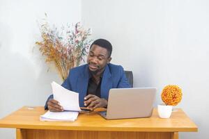 Ethnic young man reading document and writing in planner while sitting at table and working on remote project at home photo