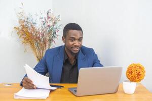 Ethnic young man reading document and writing in planner while sitting at table and working on remote project at home photo