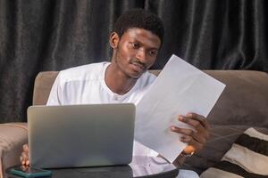 Working with papers. Serious african male entrepreneur work at home office alone hold read document hardcopy think on conditions terms. Focused young man engaged in paperwork edit text of agreement photo