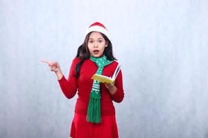 Charming young woman wearing red sweater red sweater with Santa hat and scarf, surprised to camera opening book while pointing to right, celebrating merry christmas isolated on white background photo