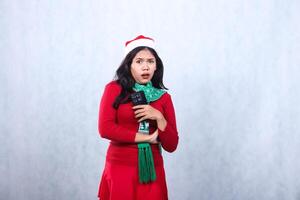 portrait of Asian girl wearing christmas sweater, Santa hat and scarf, scared shocked to camera, hands together while holding cellphone mobile phone, isolated on white background photo