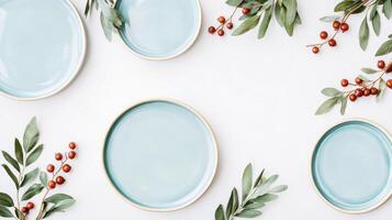 Olive green leaves and red-brown dried berries arranged around light blue ceramic plates on a natural linen surface creating a serene flat lay composition photo