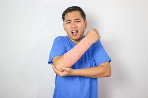 A resilient male with broken arm maintains smile while wearing splint for treatment, coping with discomfort. An upbeat Asian man in an arm sling on white background, symbolizing injury recovery. photo