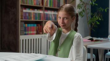 Smiling happy glad Caucasian little girl school child kid pupil joyful schoolgirl student primary education learner pointing index finger at camera inviting welcoming hand gesture calling in classroom video