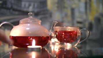 a woman's hand adjusts candles on a table cups of red tea and glass teapot sit on a stand, warmed by a candle underneath. One cup is lifted to take a sip. video