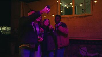 Friends celebrating at night under string lights while taking selfies with drinks in a cozy outdoor setting video