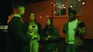 Friends enjoying a cozy evening gathering with drinks and laughter under string lights at a backyard party in fall video