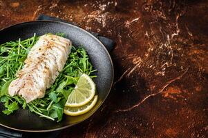Grilled cod fish fillet served with green salad in a plate. Dark background. Top view. Copy space photo