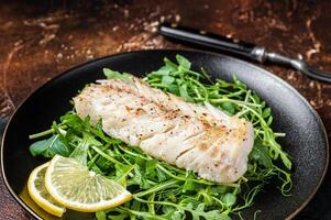 Grilled cod fish fillet served with green salad in a plate. Dark background. Top view photo