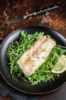 Grilled cod fish fillet served with green salad in a plate. Dark background. Top view photo